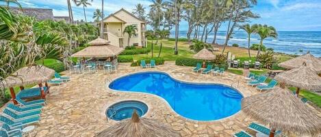A pool area with lounge chairs and thatched umbrellas, surrounded by palm trees and a view of the ocean.