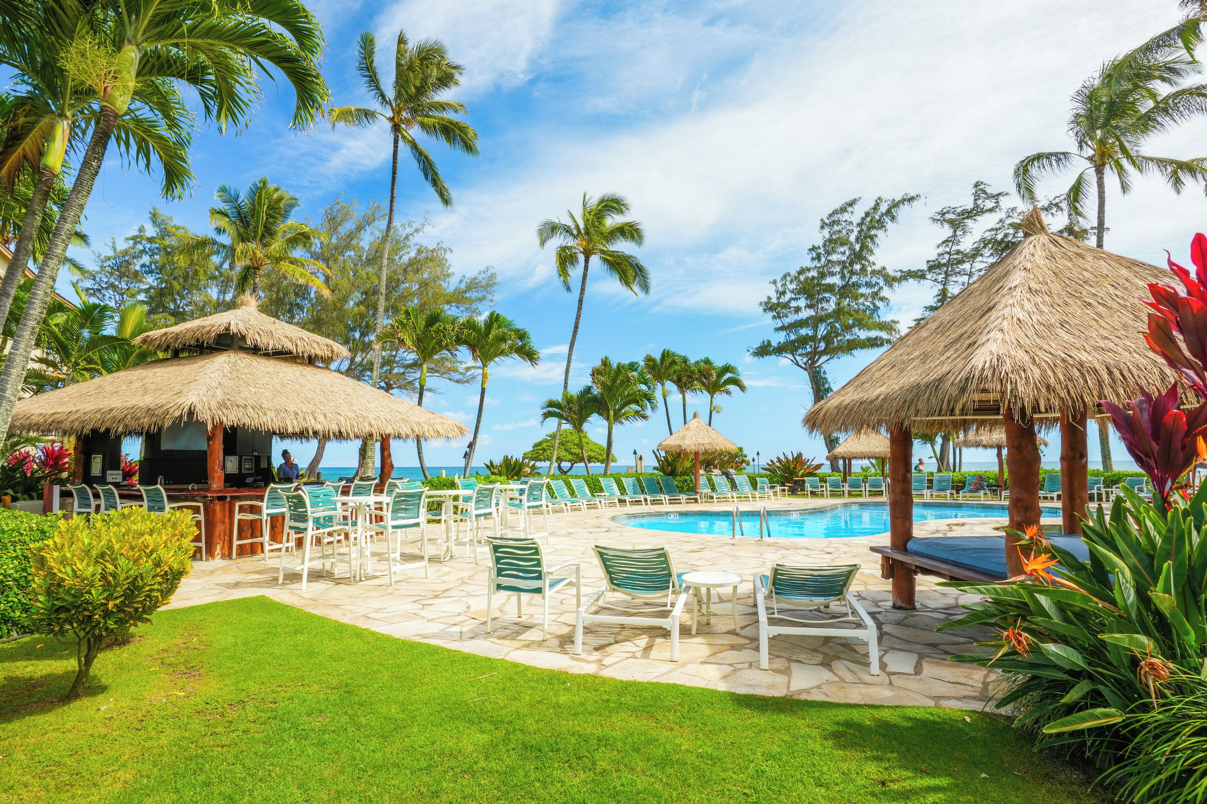 A poolside area with lounge chairs and a bar, surrounded by palm trees and a clear sky.