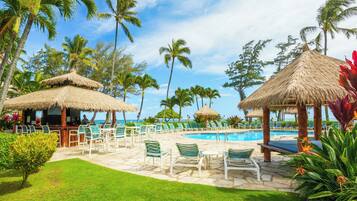 A poolside area with lounge chairs and a bar, surrounded by palm trees and a clear sky.