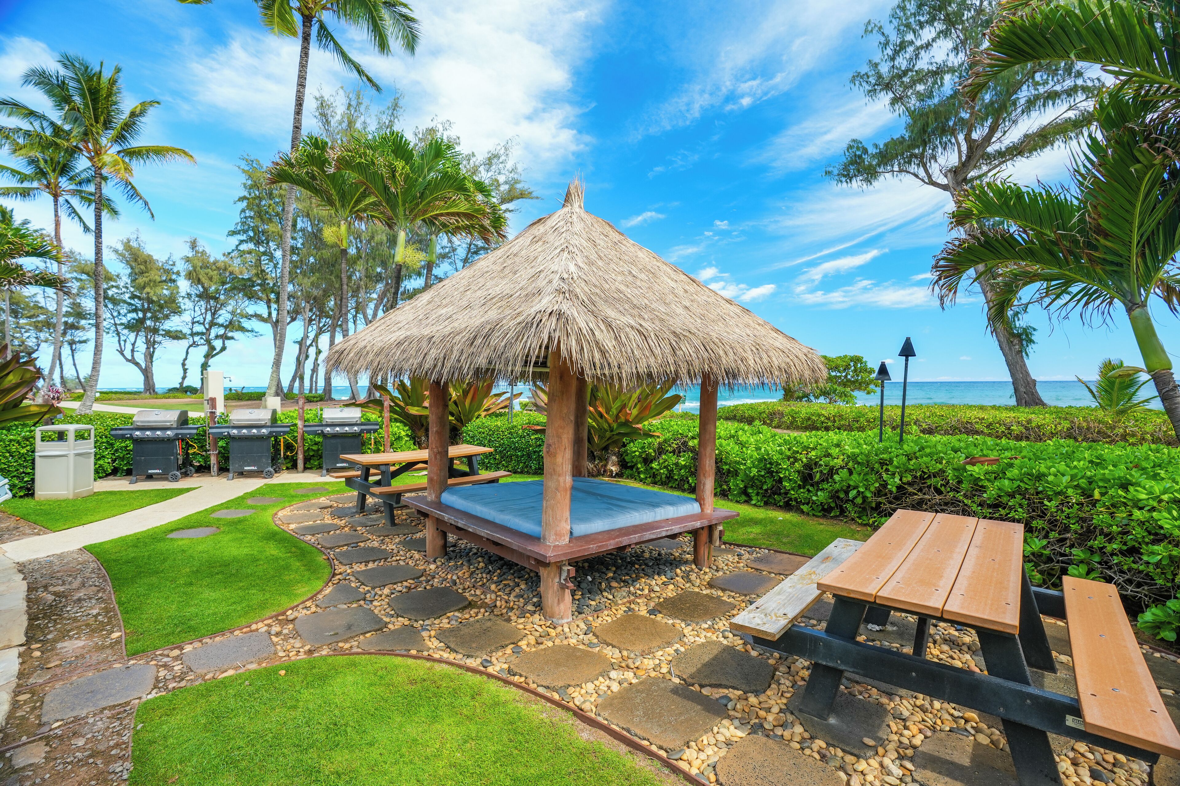 A thatched-roof gazebo with a blue cushion, surrounded by a stone pathway, palm trees, and a view of the ocean.
