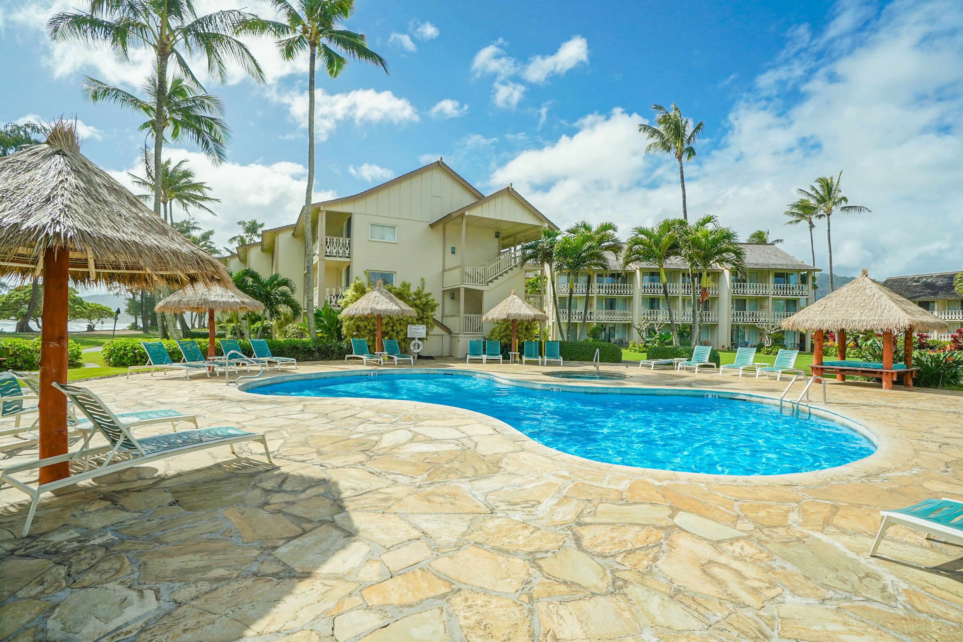A swimming pool area with lounge chairs, thatched umbrellas, and a building with balconies.