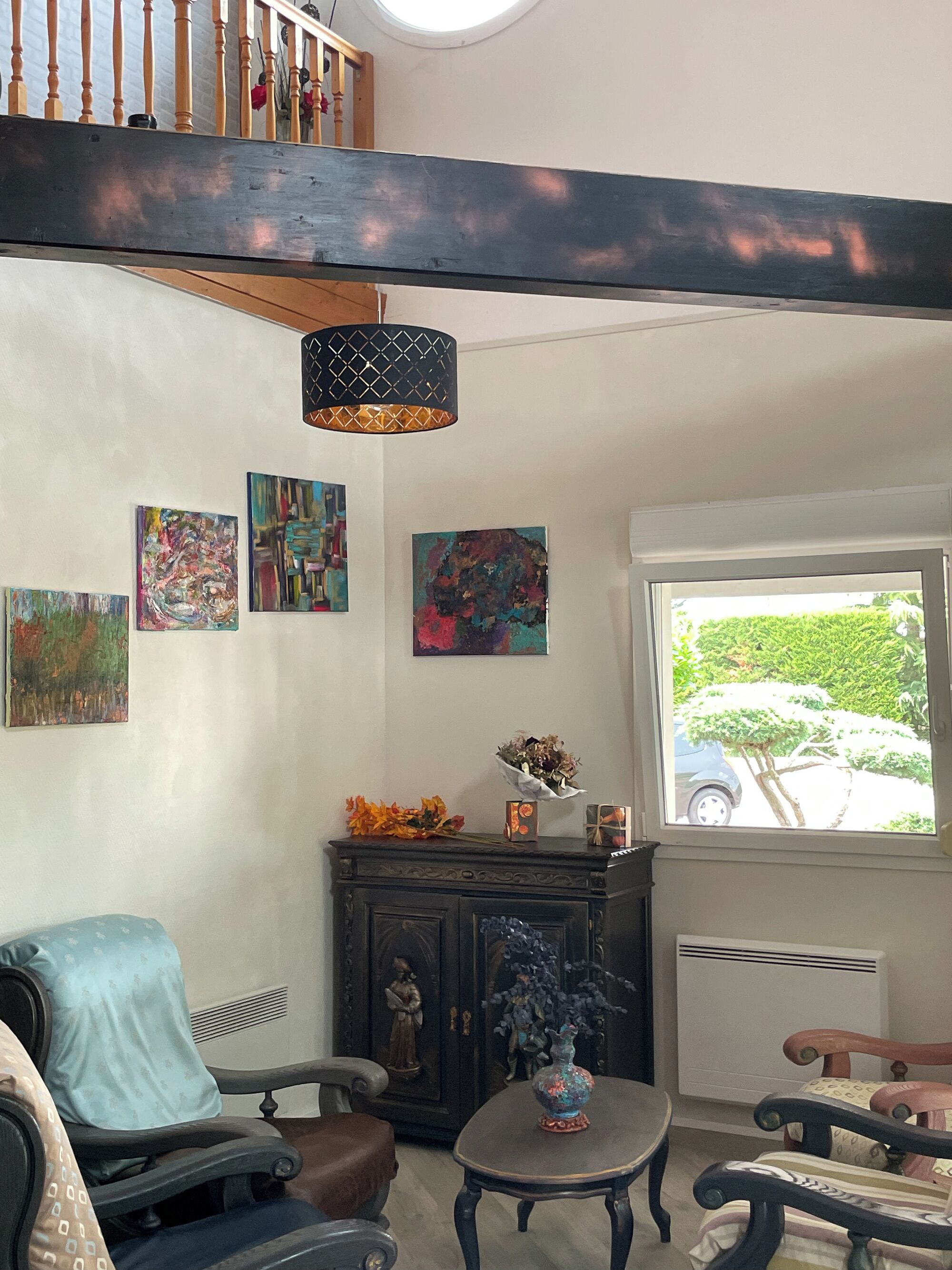 A living room with a wooden ceiling, a black pendant light, a dark wooden cabinet, and a window with a view of greenery and a parked car.