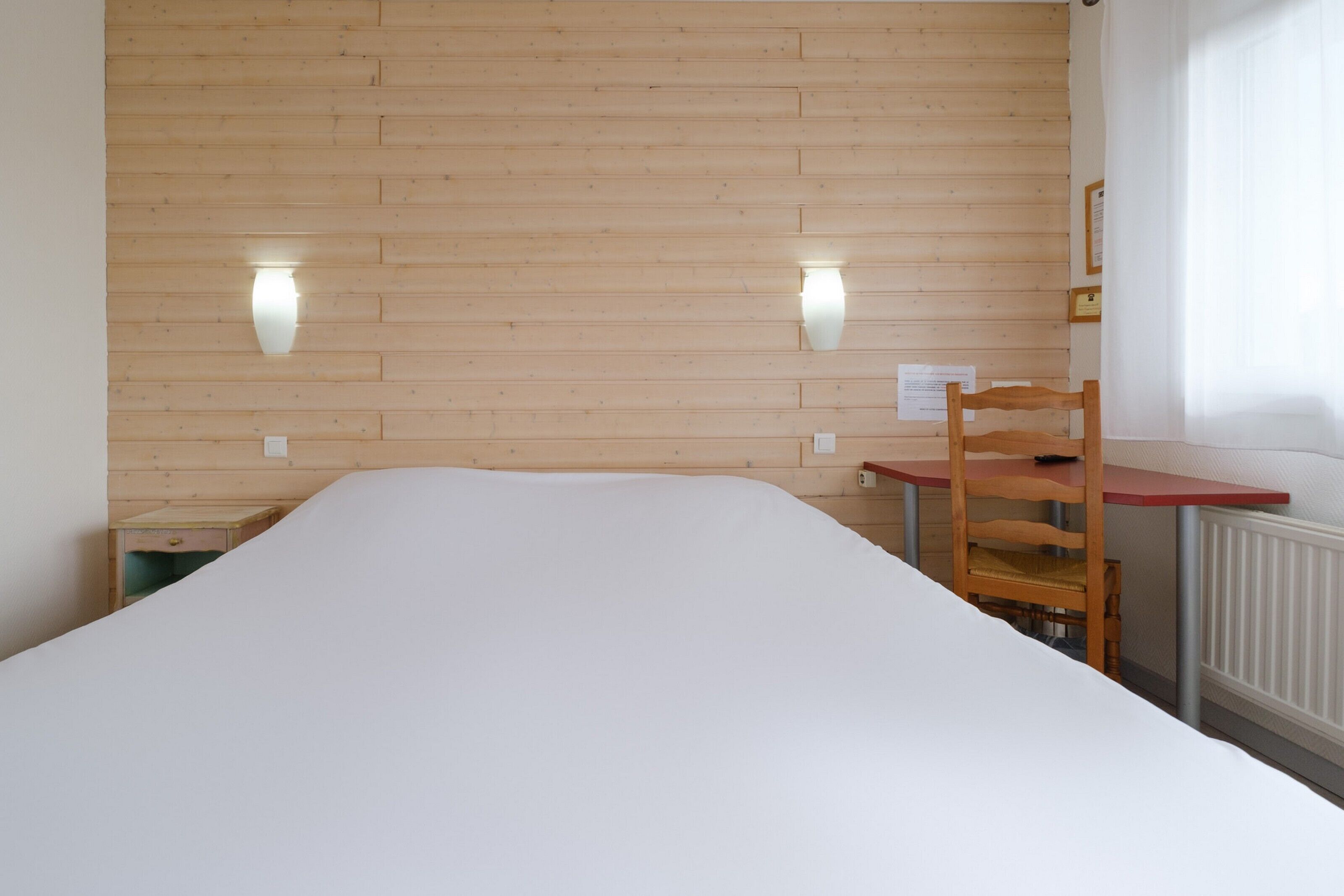 Una habitación con cabecera de madera, cama con ropa de cama blanca, una silla de madera y una pequeña mesa.