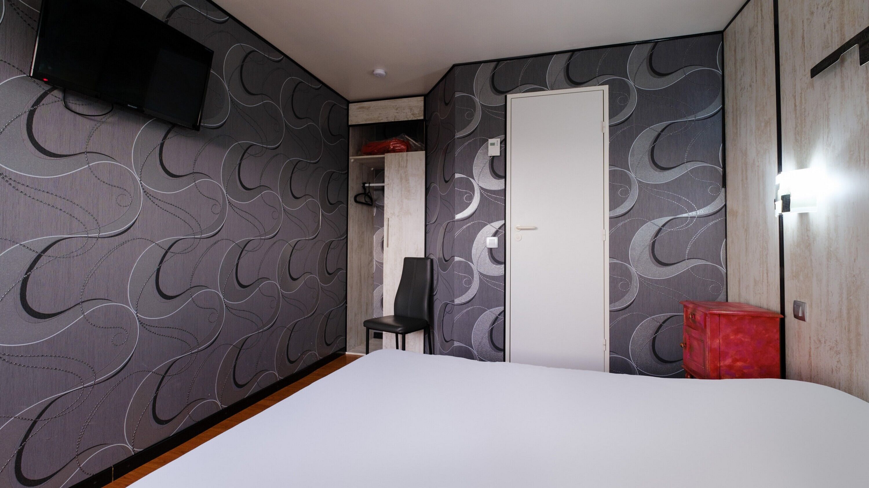 Una habitación de hotel con una cama, televisión montada en la pared, una puerta y una silla.