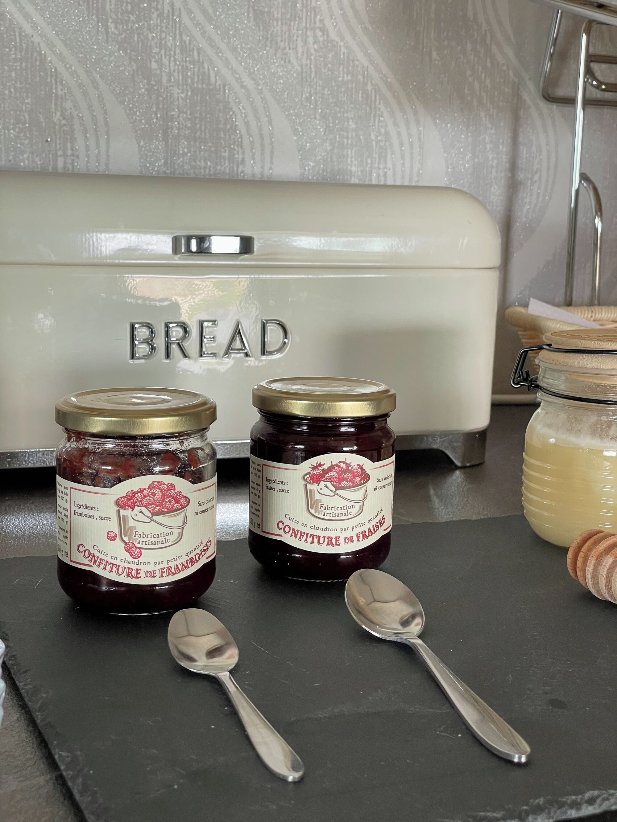 Two jars of confiture de fraises on a black surface with spoons beside them, a white appliance with the word "BREAD" on it, and a honey container in the background.