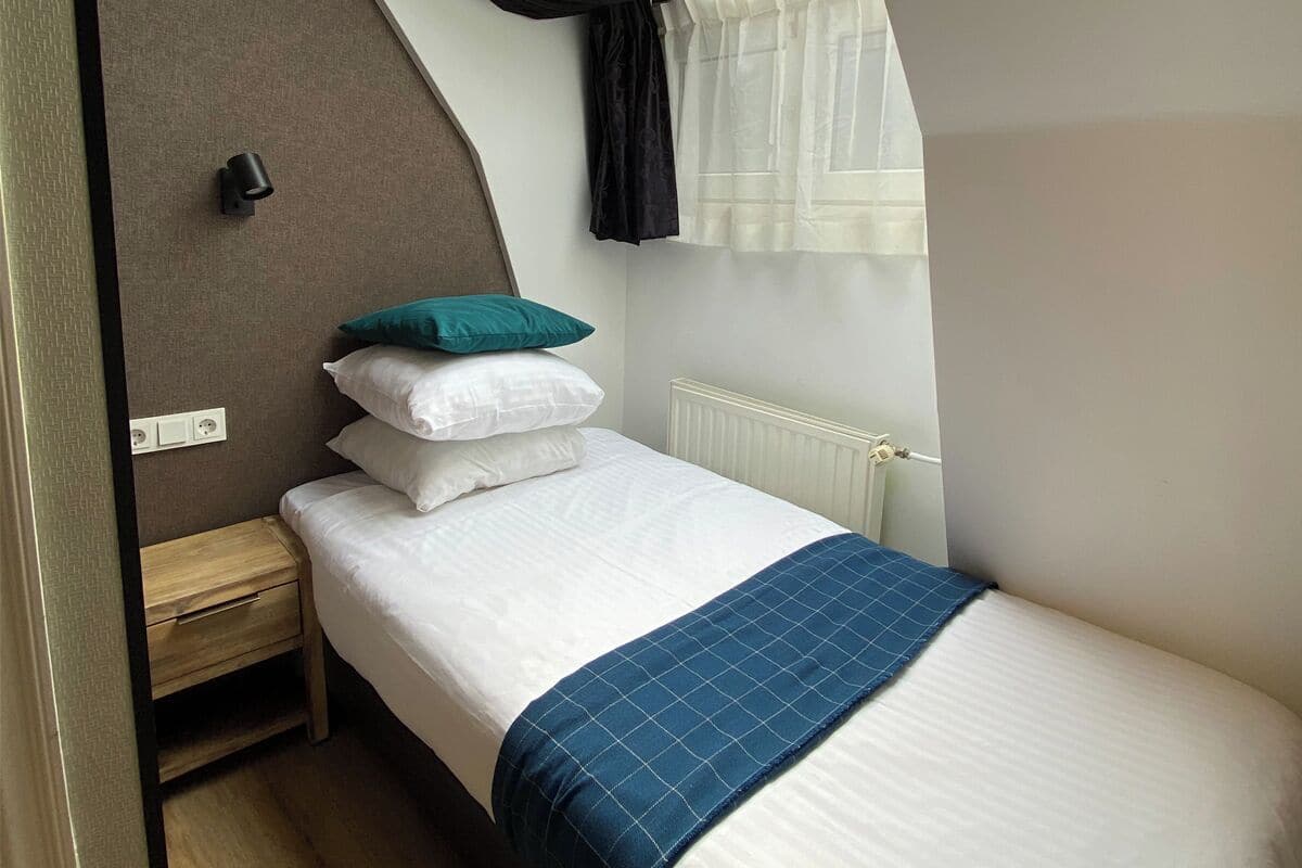 A single bed with white bedding and a blue checkered throw, a wooden nightstand, a wall-mounted light, and a window with curtains.