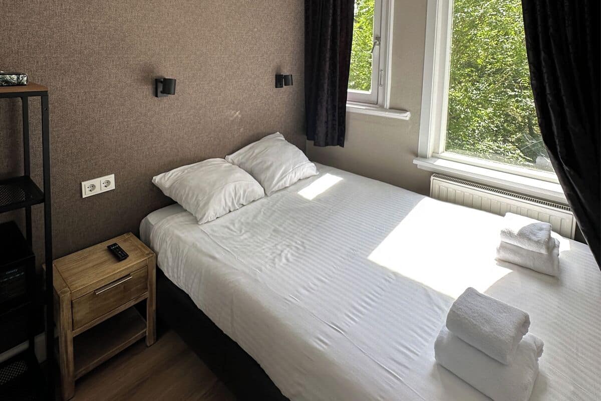 A neatly made bed with white linens, a bedside table with a remote control, and a window with curtains.