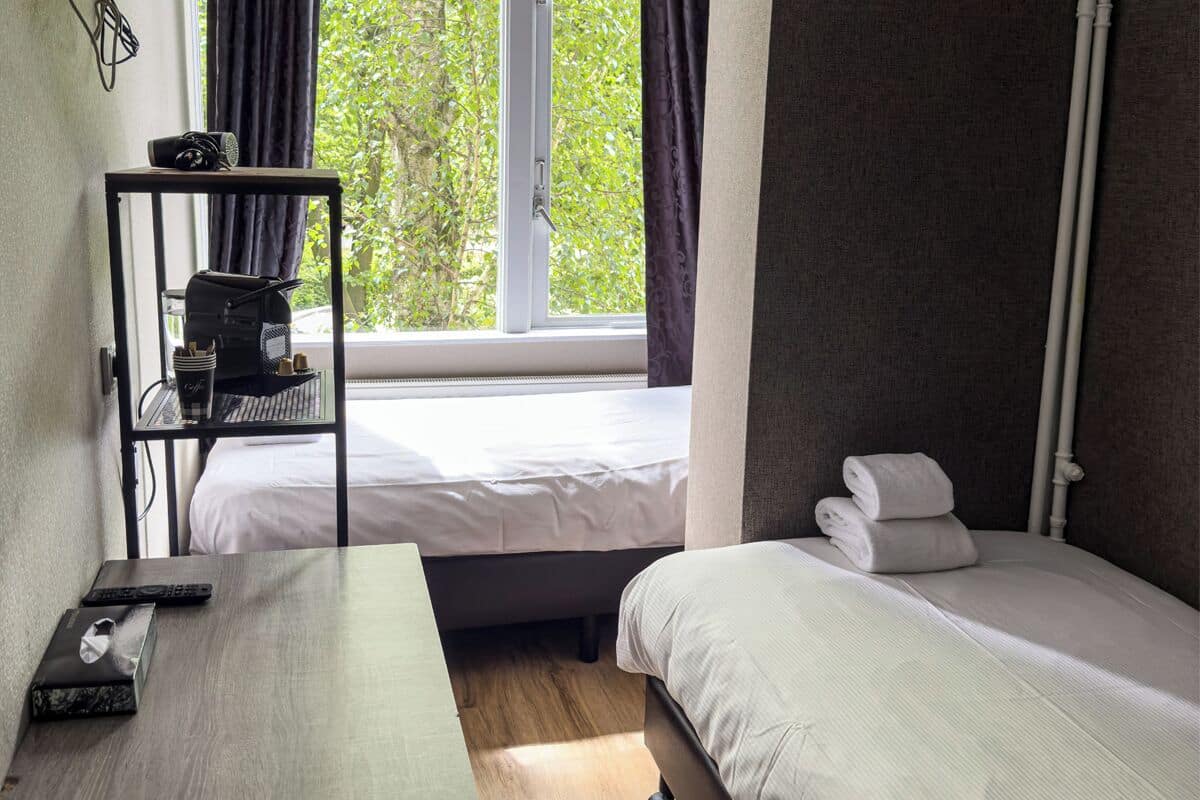 A hotel room with a bed, a small table, a shelf with items, and a window with curtains.