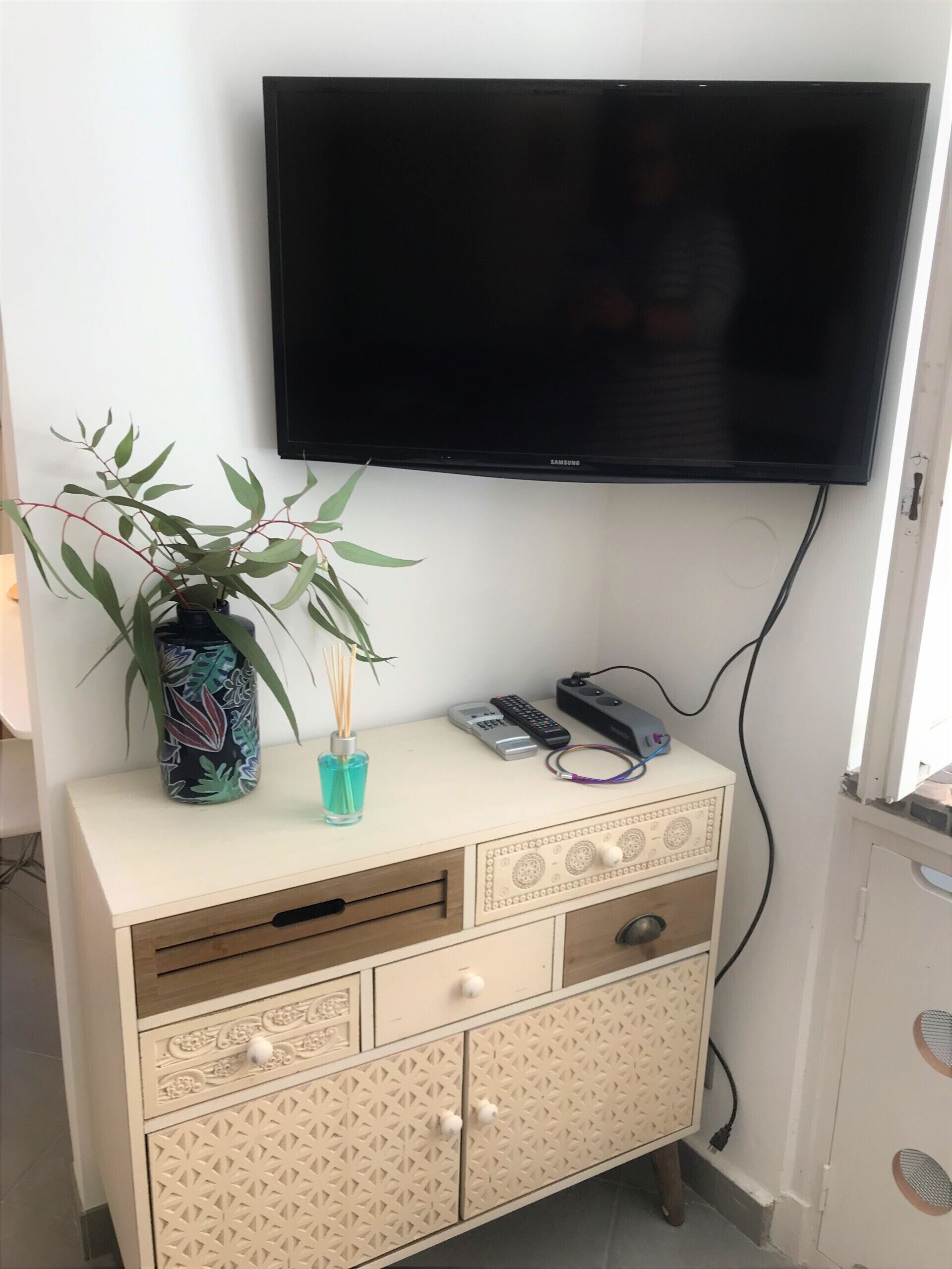 Et fjernsyn hængt på væggen over et skab med skuffer og en vase med en plante.