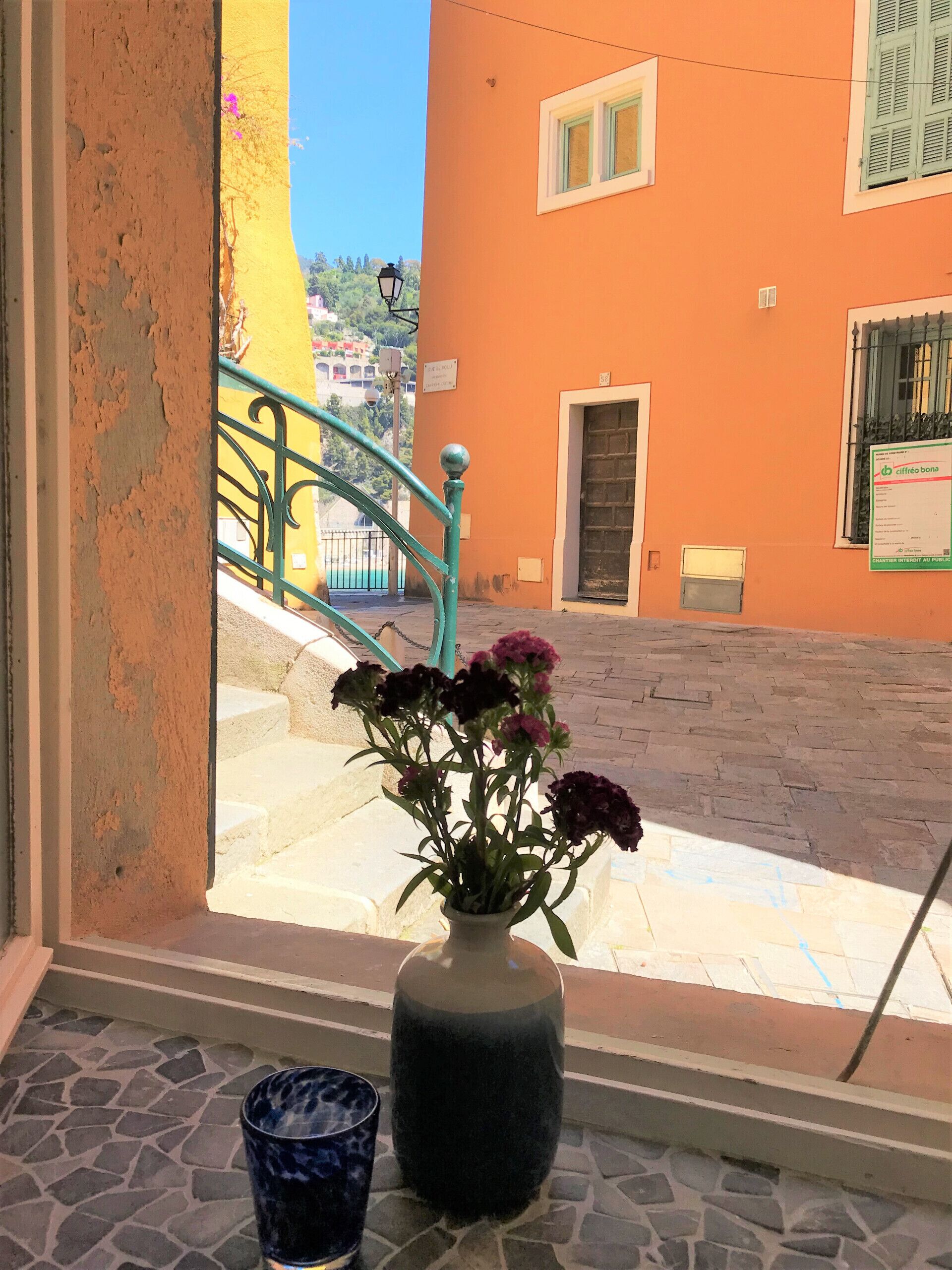 En utsikt genom ett fönster mot en byggnad, med en vas med blommor på fönsterbrädan och ett blått glas på golvet.