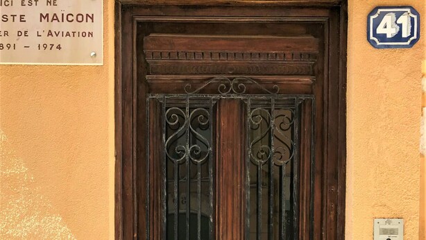 A wooden door with a metal gate, a plaque on the wall, and a house number sign.