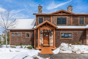 A two-story log cabin with a stone chimney, a wooden front porch, and a snow-covered roof.
