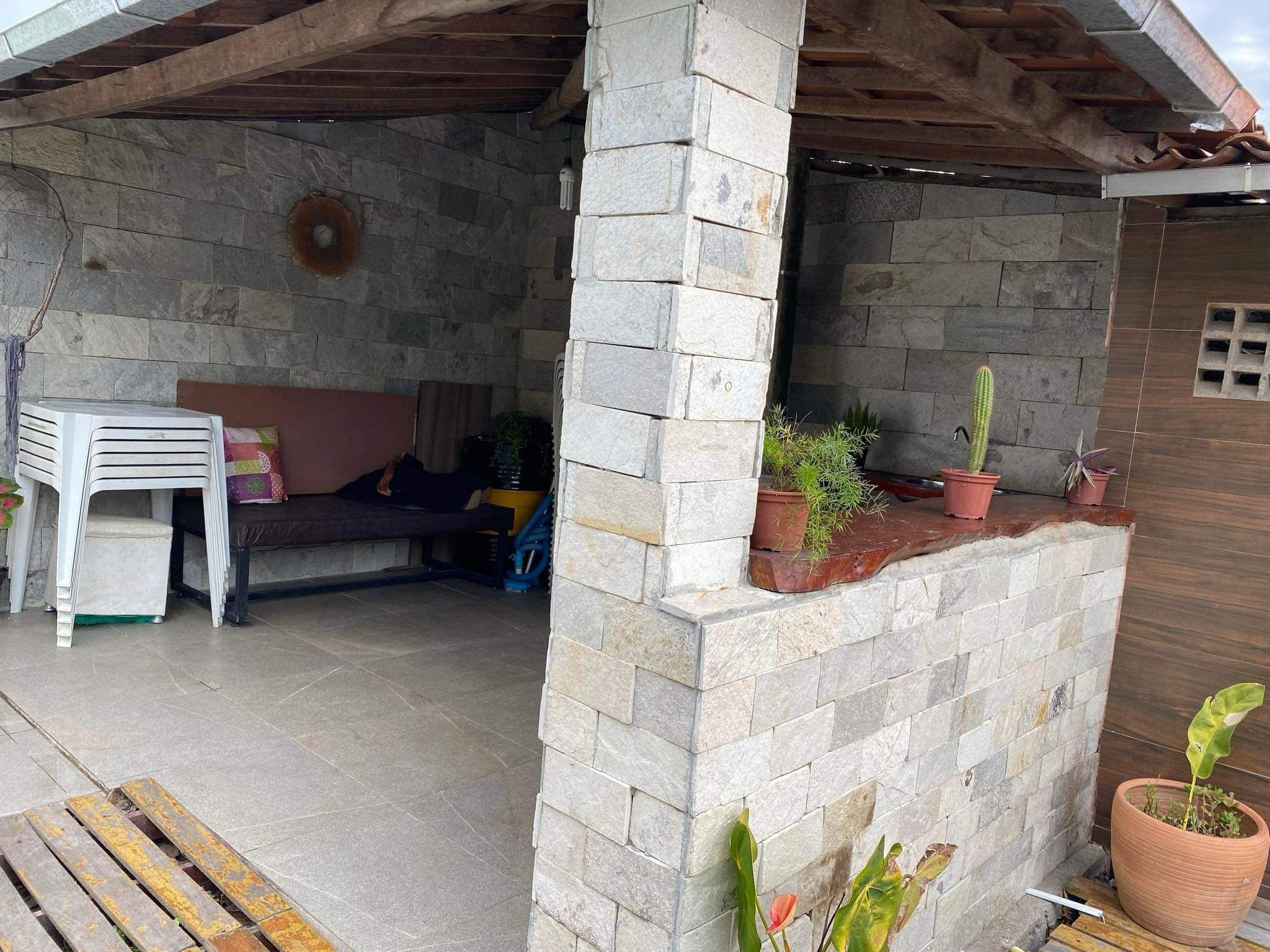 Un espacio al aire libre cubierto con paredes de piedra, una banca, plantas en macetas y una silla de plástico blanco.