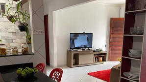 Een moderne woonkamer met een televisie, een eethoek met een zwarte tafel, rode stoelen en een rood vloerkleed. Er zijn planken met servies en een plant aan de muur.