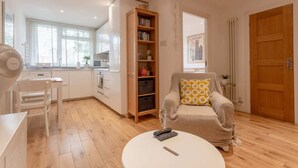 Una sala moderna con una mesa de centro redonda blanca, un sofá color beige con un cojín estampado, una estantería de madera y una zona de cocina con armarios blancos y una ventana.