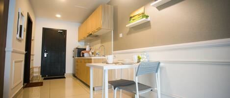 A modern kitchen with white cabinets, a white table, and a chair. There is a black door and a shelf with decorative items.