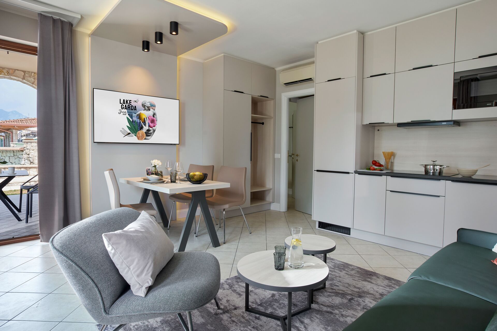 Un salon moderne avec un canapé gris, une petite table ronde et un coin repas comprenant une table et des chaises blanches. On peut apercevoir une cuisine avec des meubles blancs et un fourneau noir.