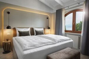 A modern hotel room with a large bed, two bedside lamps, a view of the outdoors through a window, and a small round table with a lamp.