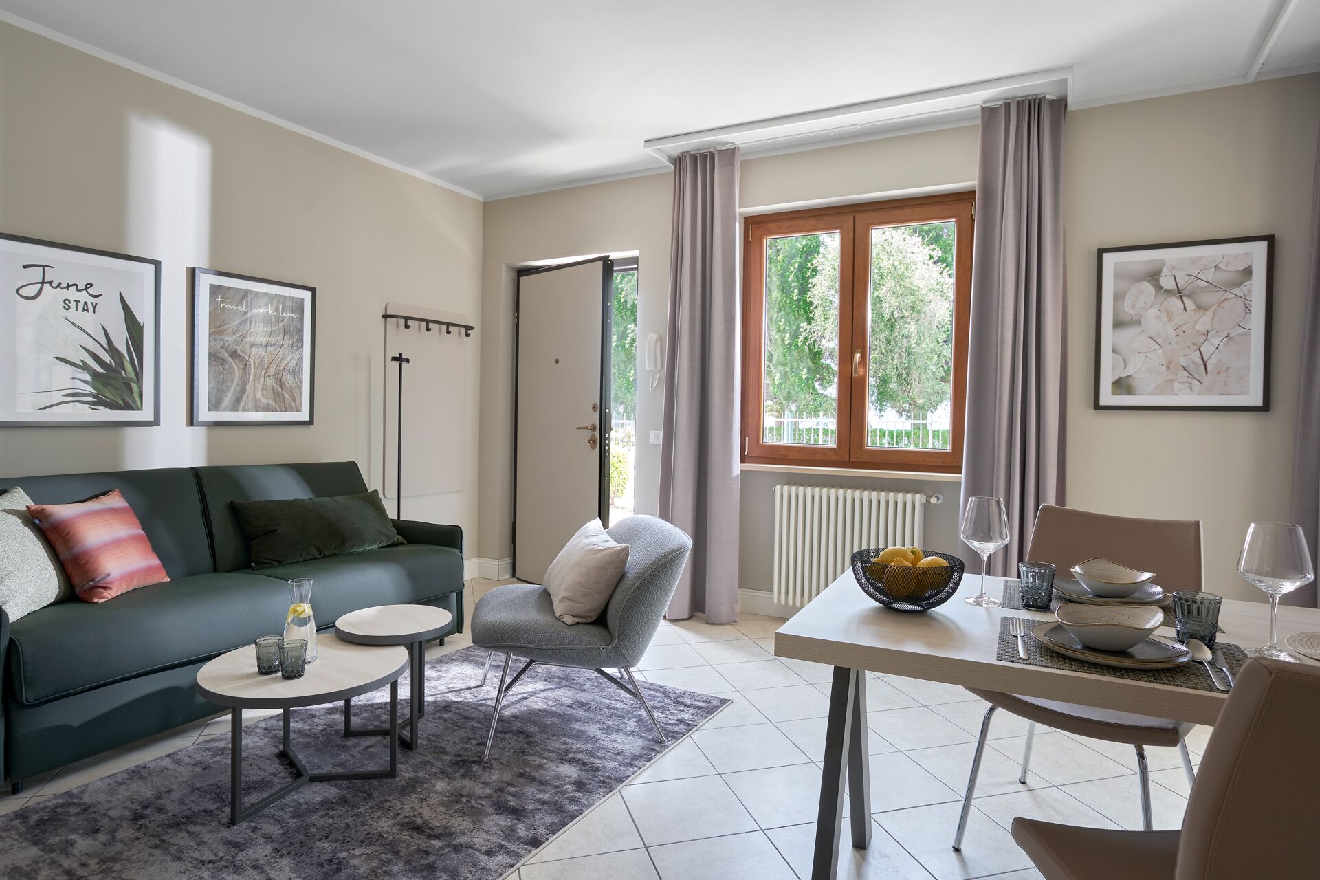 Ein modernes Wohnzimmer mit einem grünen Sofa, einem grauen Sessel, einem Esstisch mit einer Zitronenschale und einem Fenster mit Vorhängen.