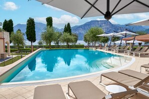 A swimming pool with lounge chairs and umbrellas, surrounded by trees and mountains in the background. - June Stay Lake Garda (Brenzone sul Garda)