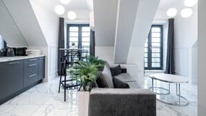 A modern kitchen with dark cabinets, a marble countertop, and a coffee machine. There is a dining area with a grey sofa, a small round table, and a potted plant. - RentPlanet - Bulwar Staromiejski (Wroclaw)