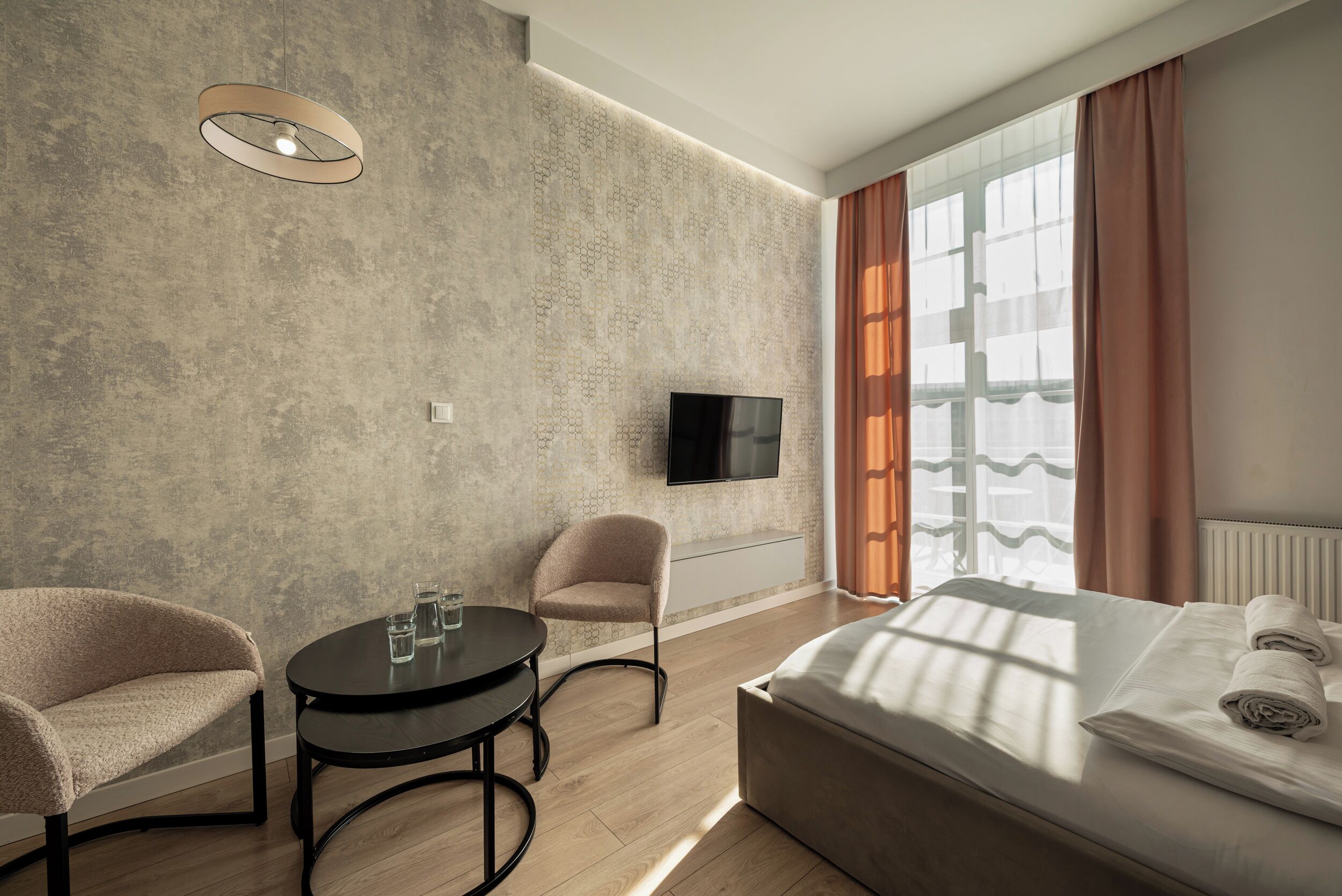 Habitació d'hotel moderna amb un llit gran, una televisió, una taula rodona amb cadires i una finestra amb cortines.