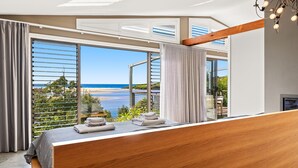 Une chambre avec une grande fenêtre donnant sur une plage, une tête de lit en bois et une vue sur l’océan.