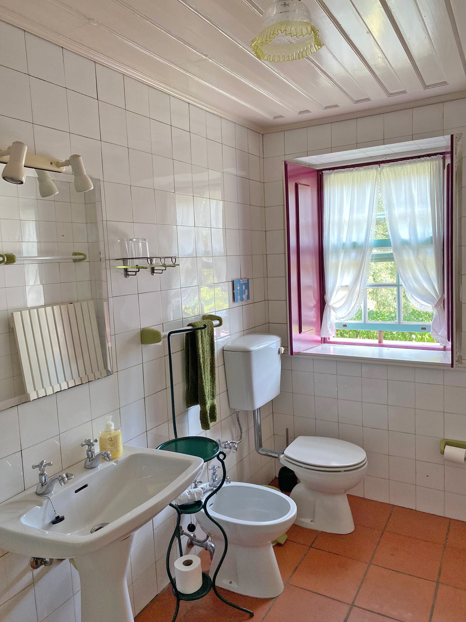 A bathroom with white tiled walls, a toilet, a bidet, a sink, a mirror, and a window with curtains.