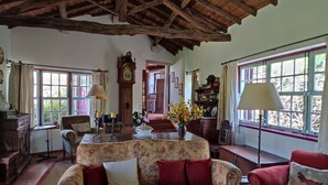 Ein gemütliches Wohnzimmer mit einem floralen Sofa, einem roten Sessel, einer Holzbalkendecke und einem großen Fenster mit Vorhängen.