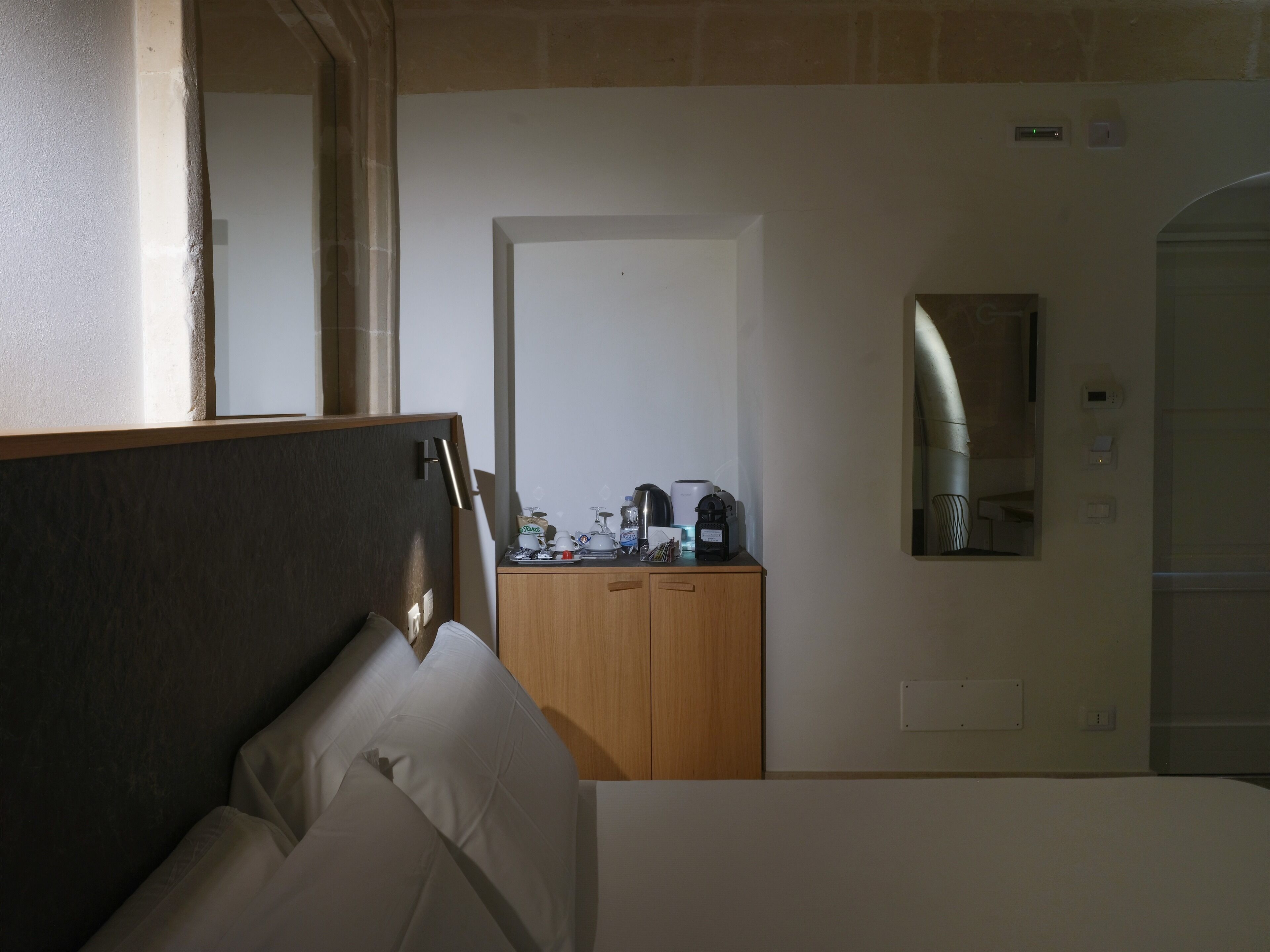 A hotel room with a bed, a wooden wardrobe, a desk with a coffee maker, and a mirror on the wall.