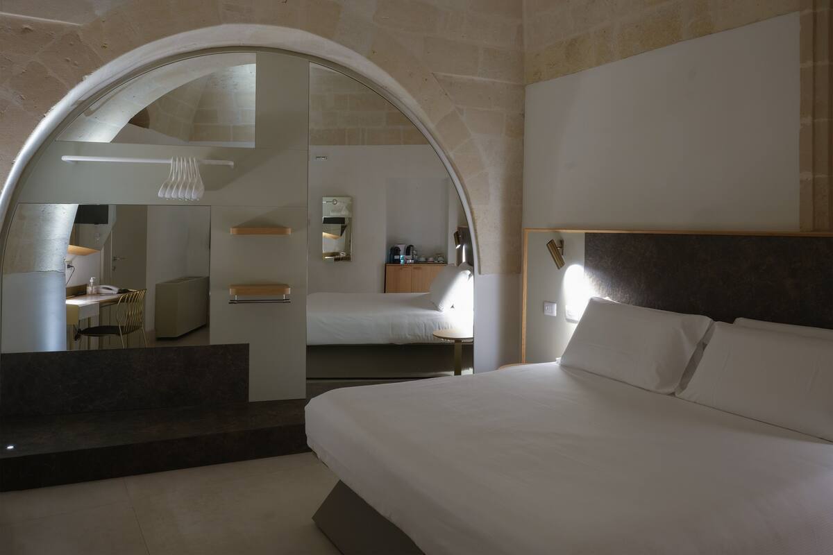 A modern hotel room with a large bed, a desk with a chair, a mirror, and a small seating area.