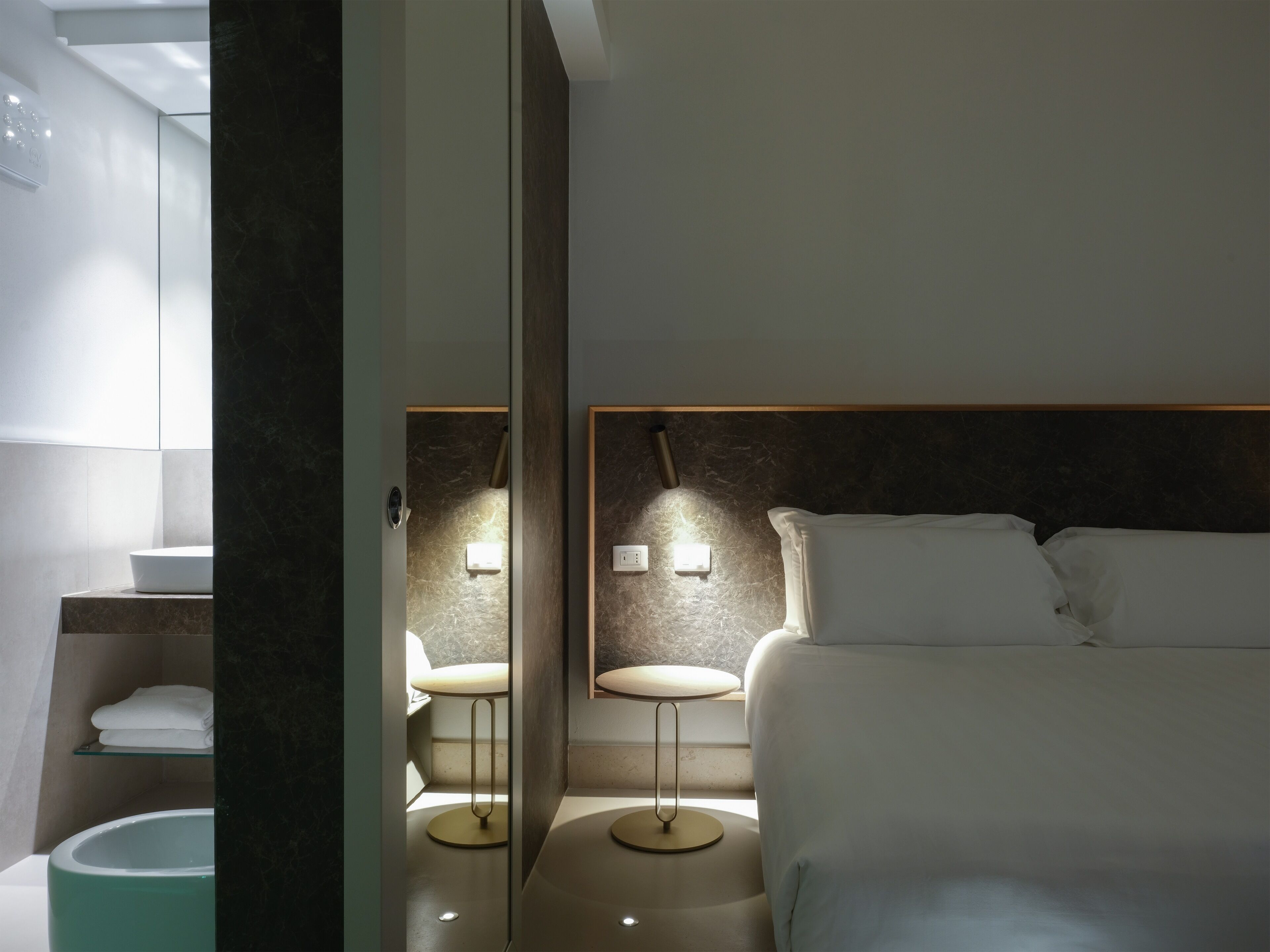 A modern hotel room with a bed, bedside tables, a small round table, and a built-in bench.