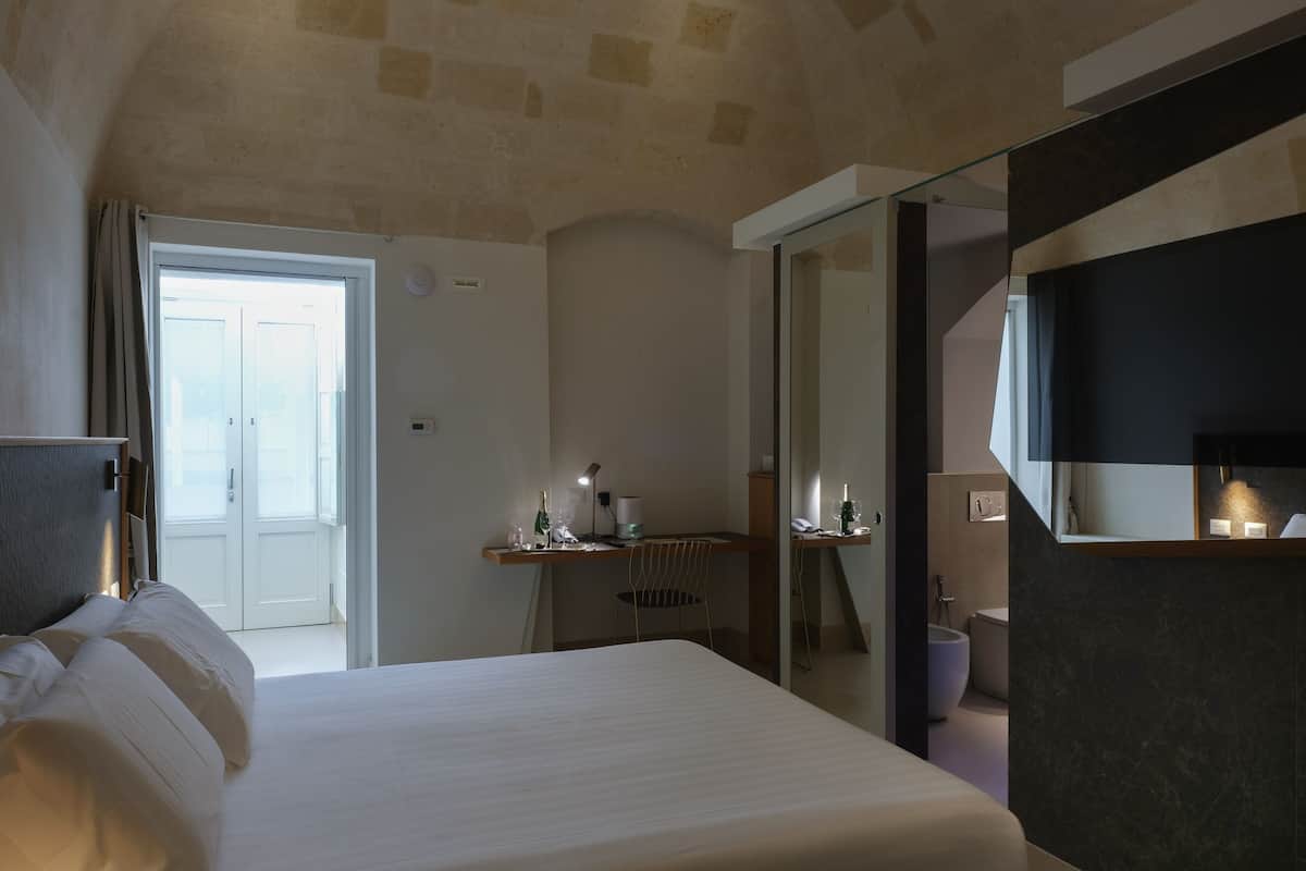 A hotel room with a bed, a desk with a lamp, a mirror, and a door leading to a balcony.