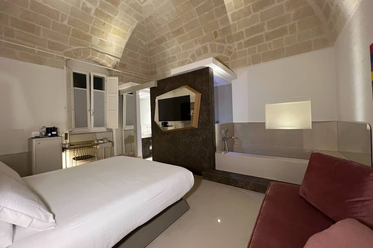 A bedroom with a bed, a desk, a mirror, a TV, and a bathroom with a bathtub.