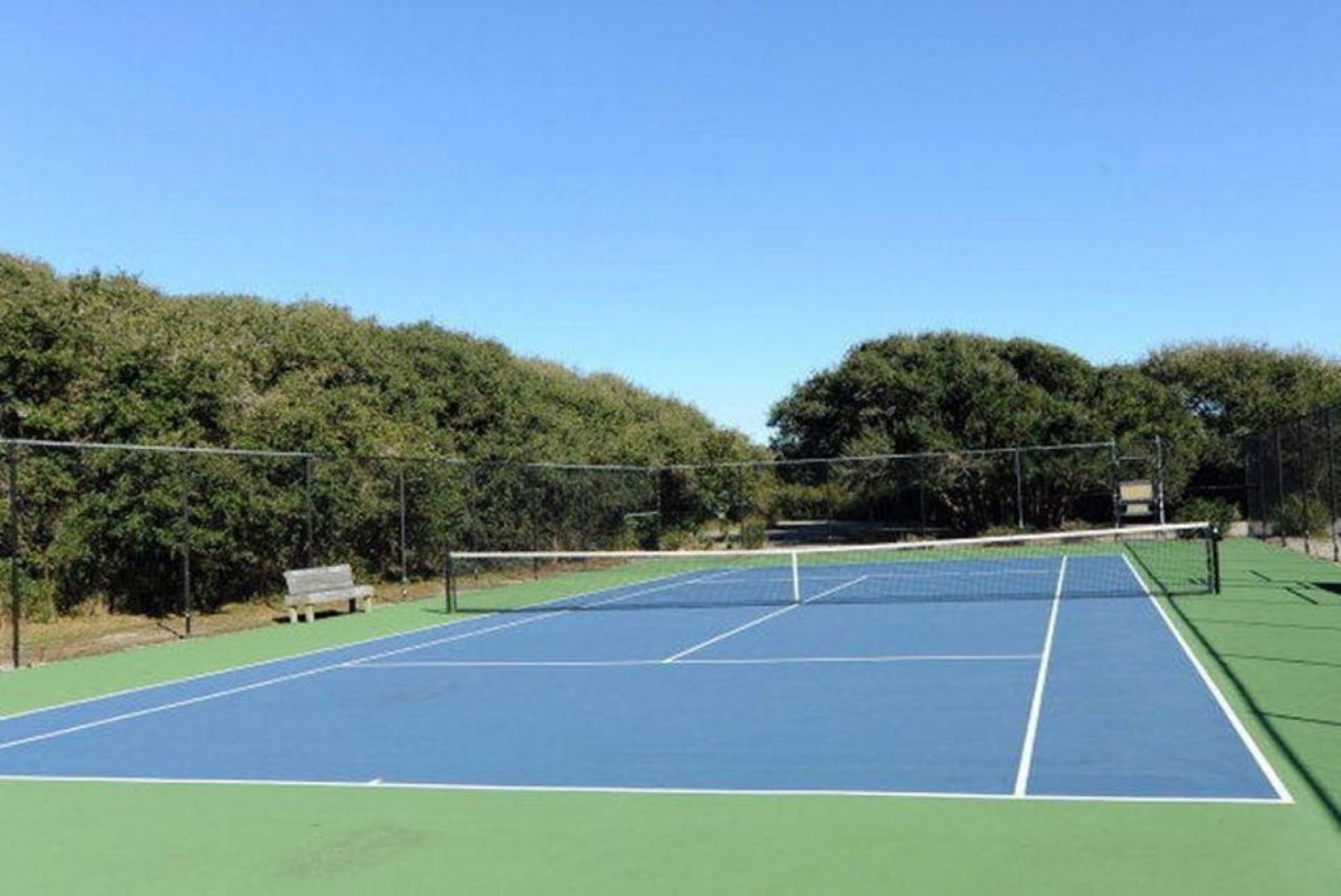 An outdoor tennis court with a net, surrounded by trees and a clear sky.