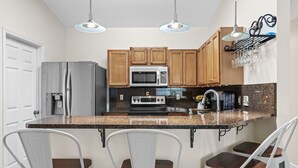 A kitchen with wooden cabinets, a stainless steel refrigerator, a microwave, and a granite countertop. There are three white bar stools with wooden seats.