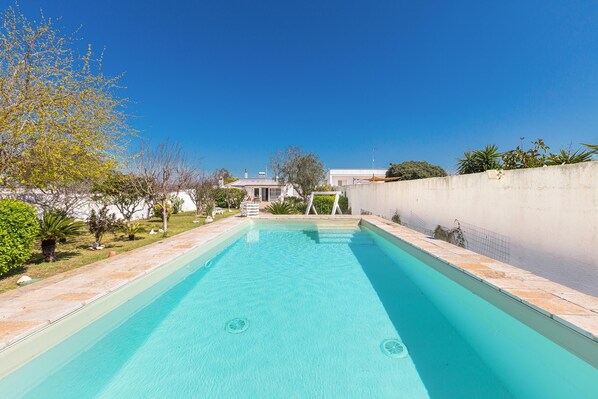 A rectangular swimming pool with clear blue water, surrounded by a low stone wall and a white fence.