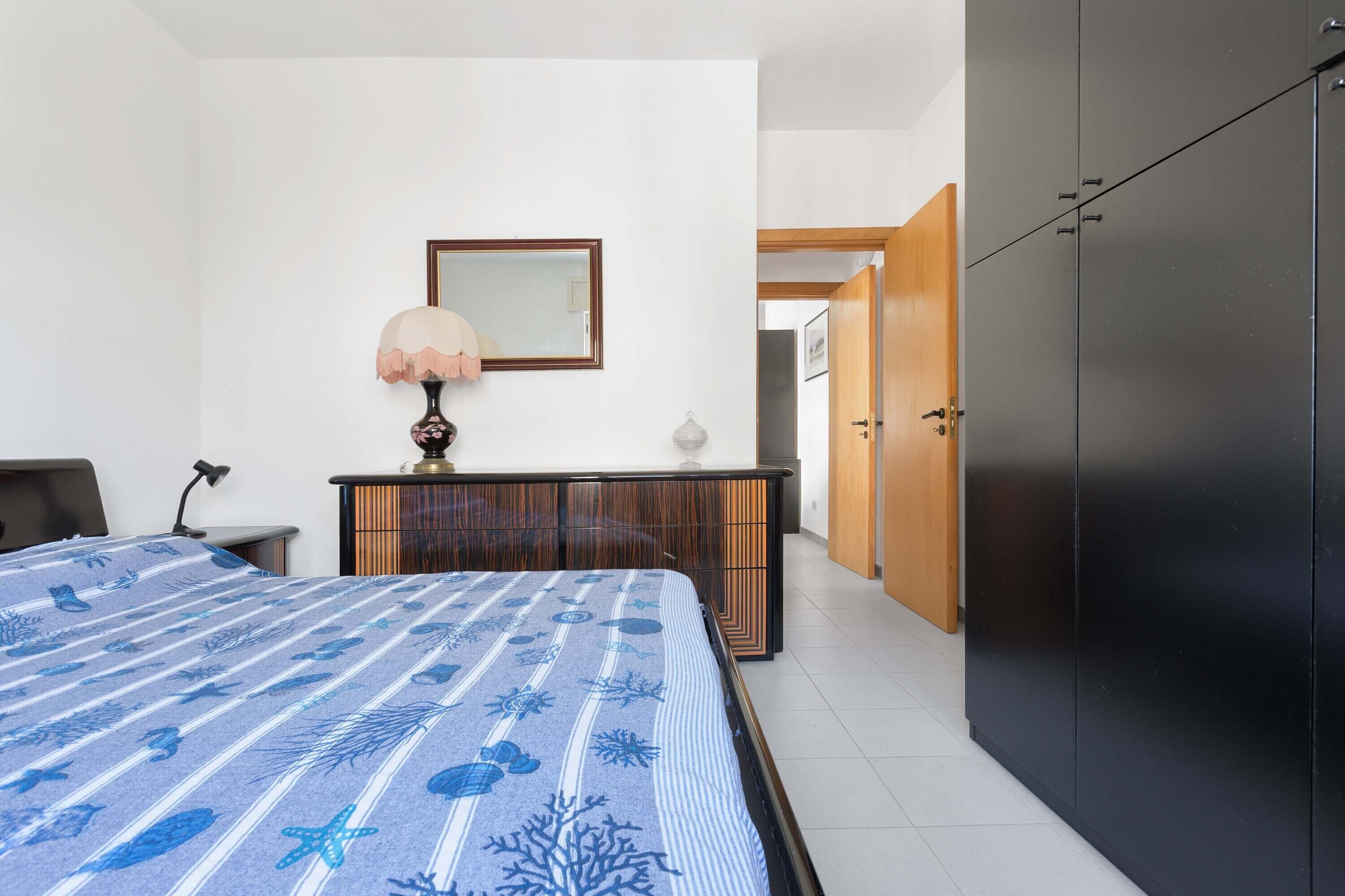 A bedroom with a bed, bedside table, lamp, dresser, wardrobe, and a door leading to another room.