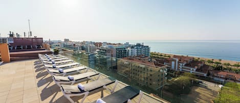 A rooftop terrace with sun loungers and a view of the sea and cityscape.