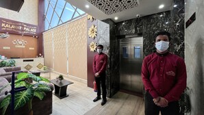 Uma área de recepção de hotel, com balcão de recepção, balcão de check-in, parede decorada com padrão floral e uma grande janela que proporciona luz natural.