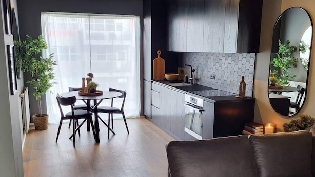 Una sala de estar moderna con un sofá marrón oscuro, un comedor con una mesa redonda y sillas, y una cocina con armarios oscuros y un salpicadero de azulejos.