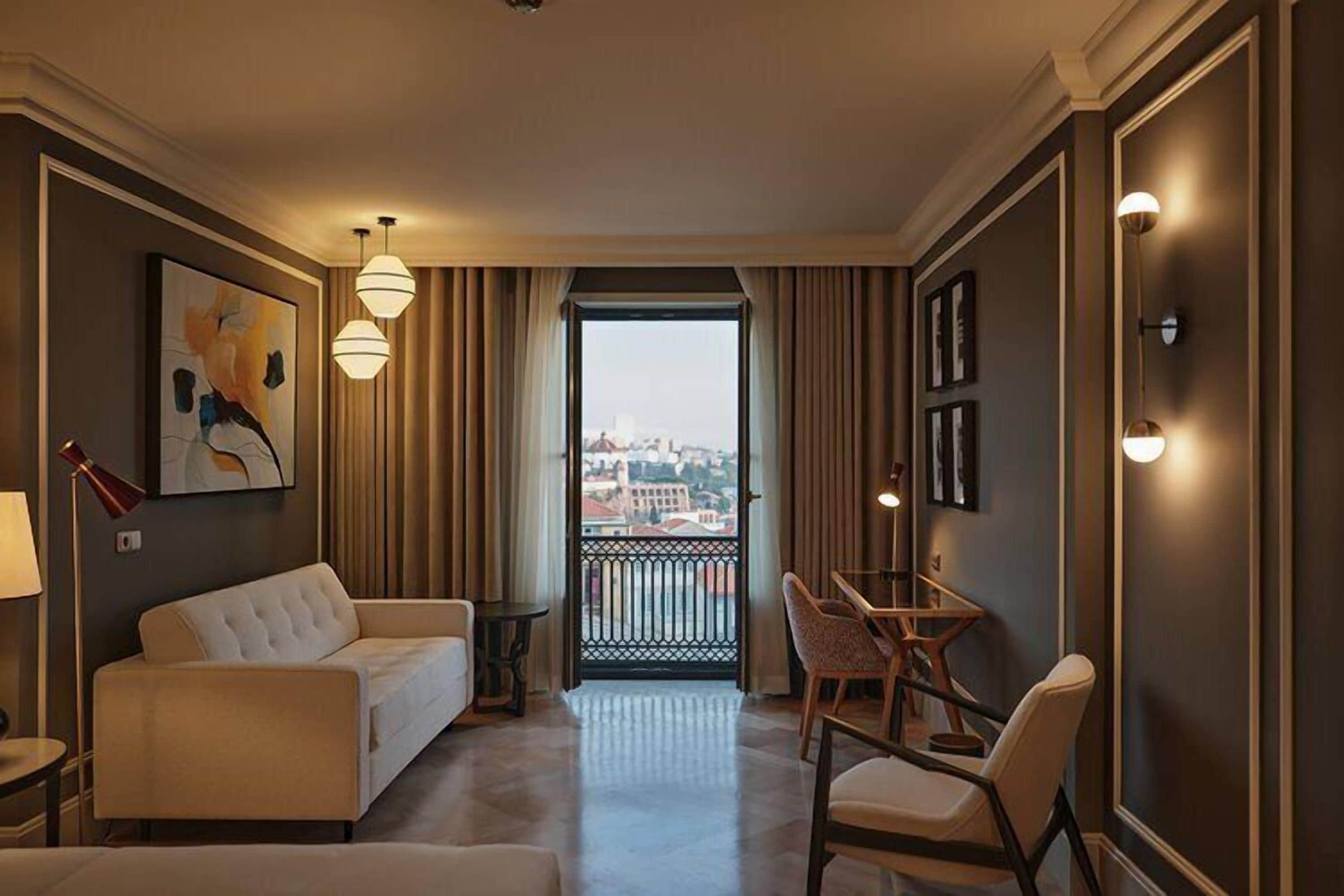A hotel room with a balcony, a sofa, a dining table with chairs, and a painting on the wall.