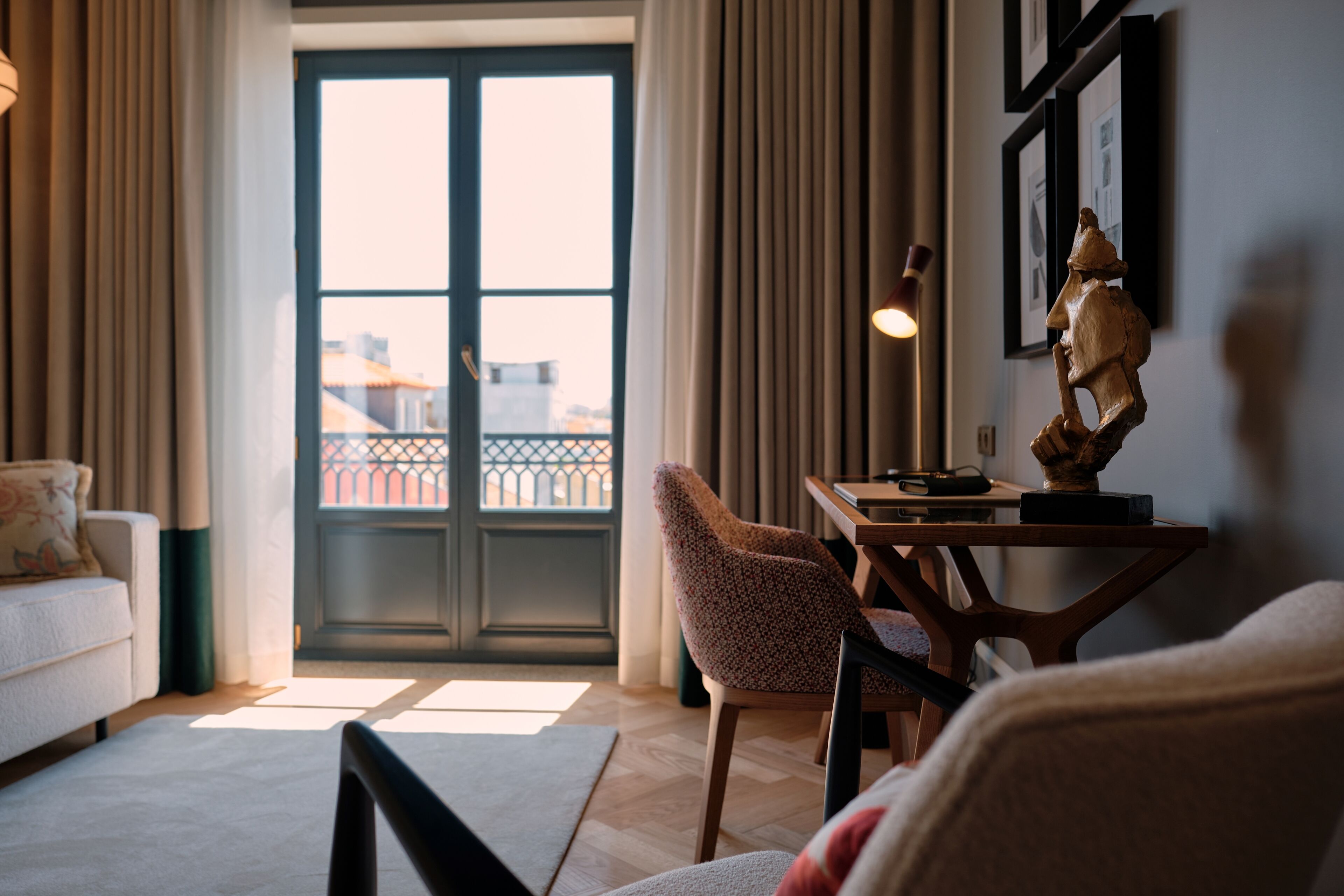 A living room with a sofa, a chair, a table with a lamp, and a sculpture. There is a door leading to a balcony with a view of buildings.