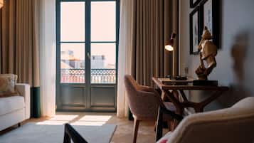 A living room with a sofa, a chair, a table with a lamp, and a sculpture. There is a door leading to a balcony with a view of buildings.