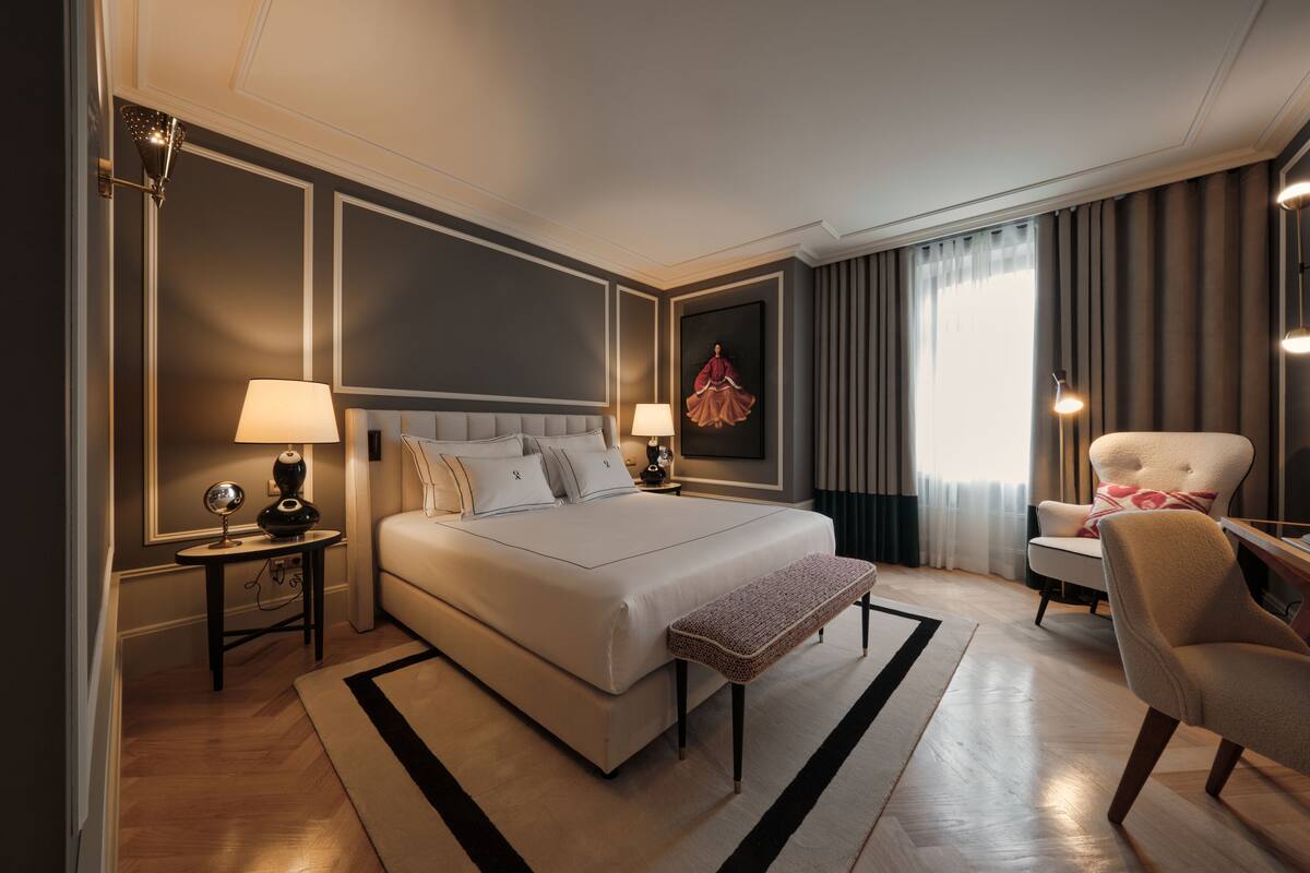A modern hotel room with a large bed, a sitting area with a chair, a desk, and a large window with curtains.