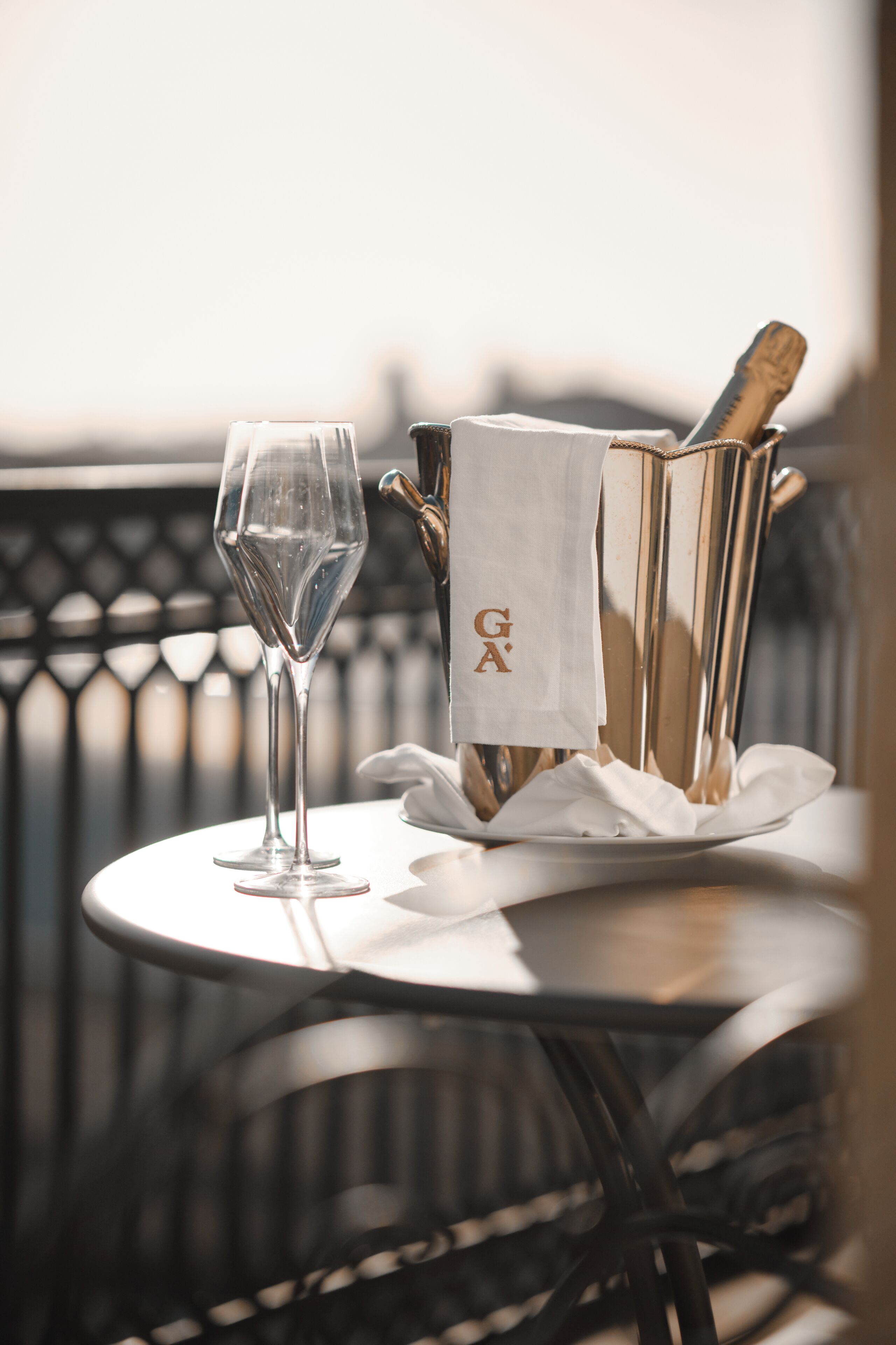 Ein Tisch mit einem Glas Wasser, einer Champagnerflasche und einem Servietten auf einer runden Metalltischplatte mit einem Stuhl.