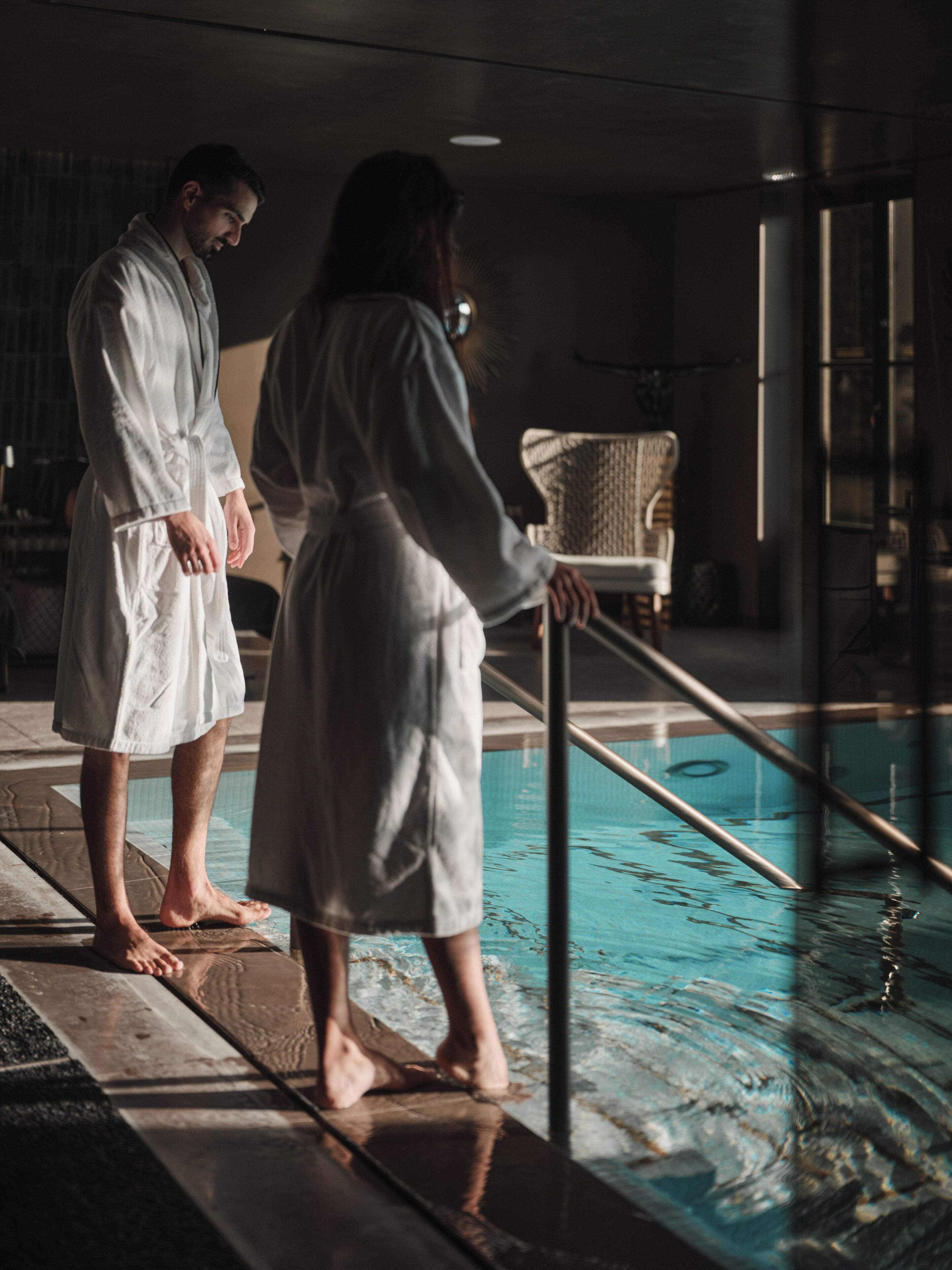 Two individuals in white robes standing by a pool with a chair and a railing in the background.