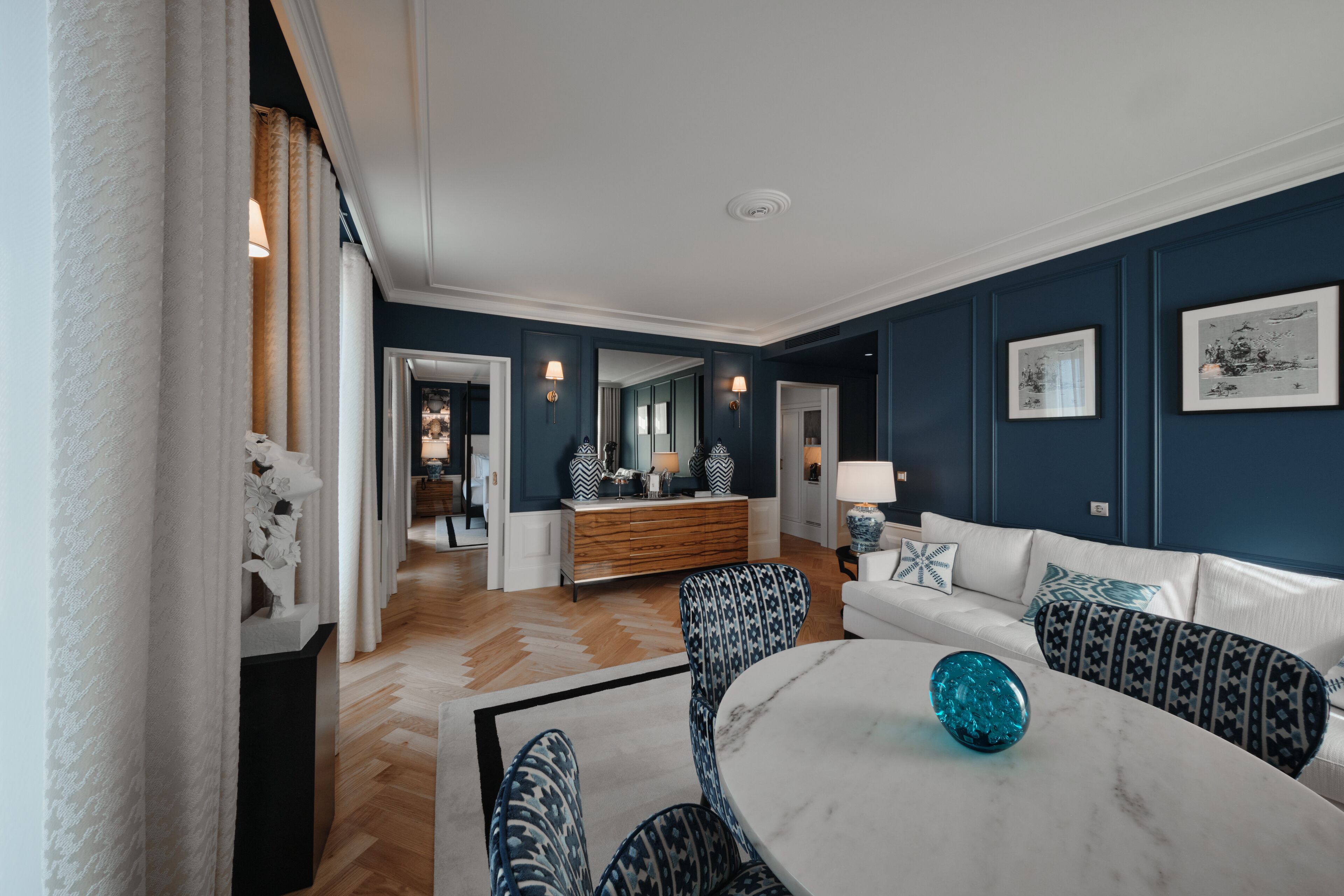 A modern living room with a white sofa, patterned armchairs, a marble coffee table, and a decorative blue glass sphere.