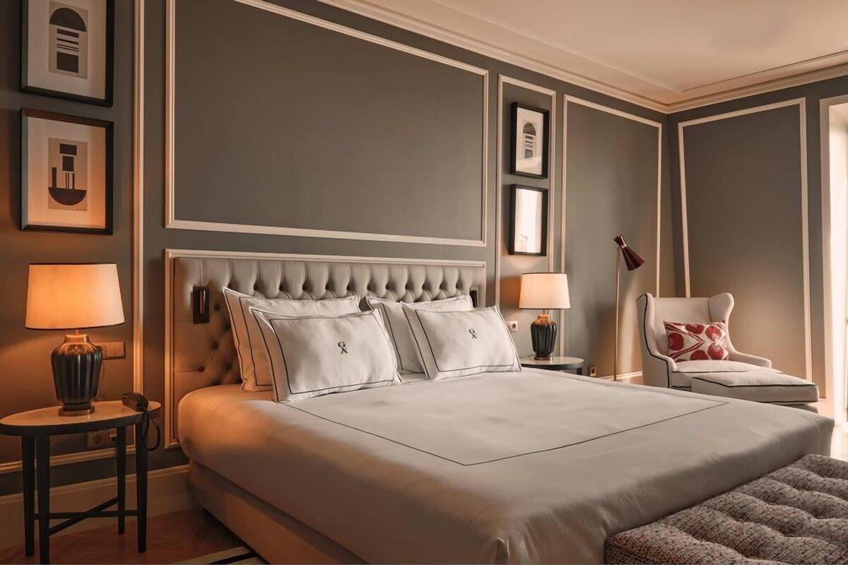 A large bed with a tufted headboard, two bedside tables with lamps, a chair with a cushion, and framed pictures on the wall.