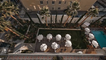 Vue aérienne d’un espace piscine aménagé sur un toit, avec des palmiers, des parasols et des chaises longues.